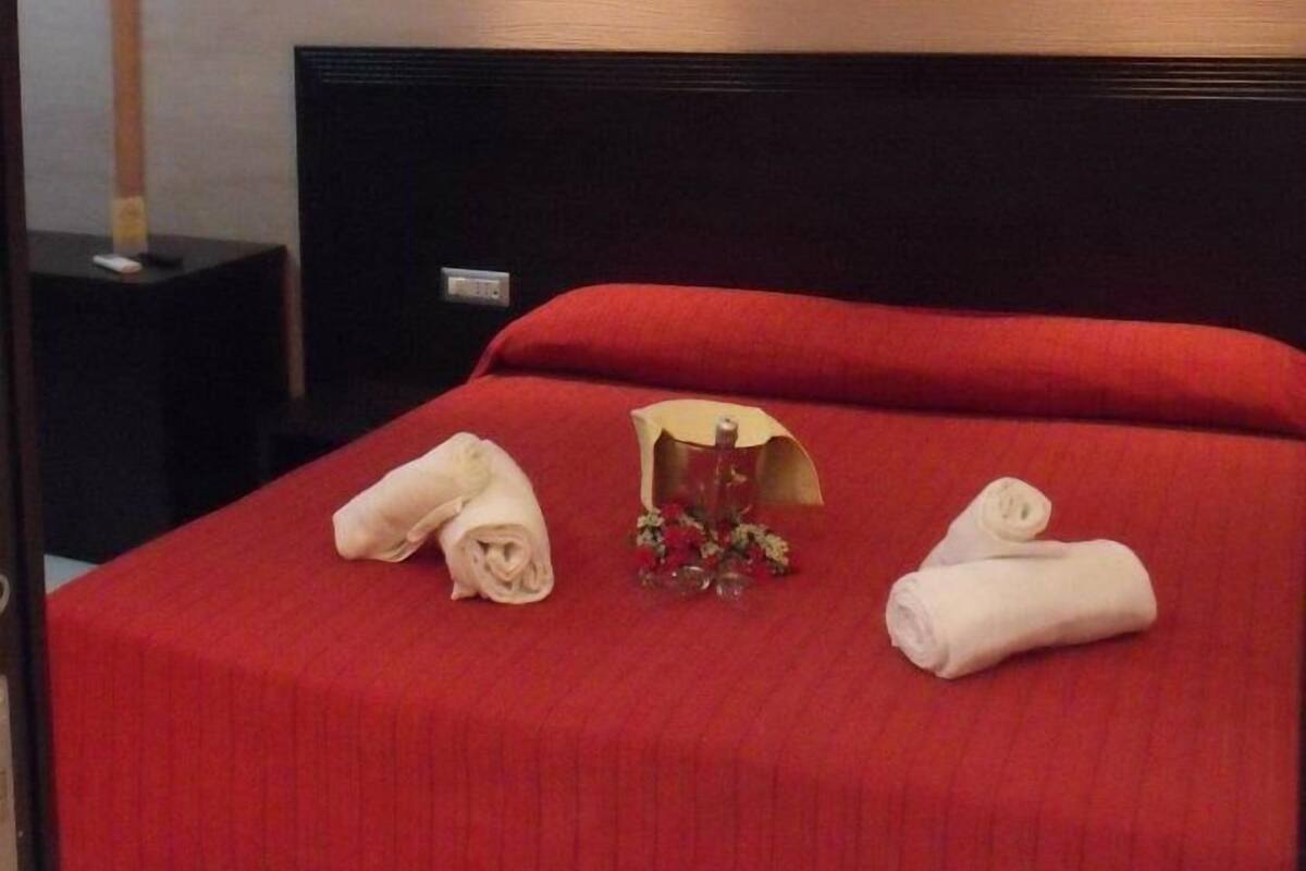 Une chambre d’hôtel avec une couvre-lit rouge, une tête de lit en bois et une petite table sur laquelle se trouve une télécommande.