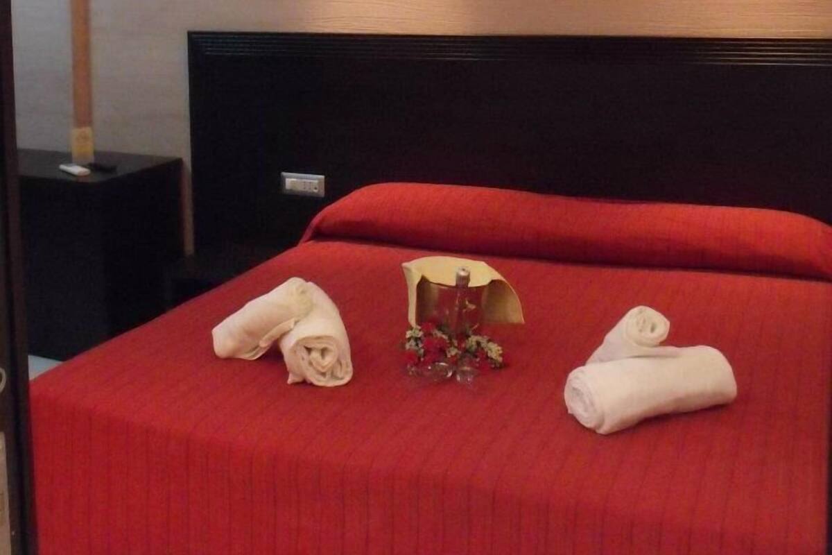 Une chambre d’hôtel avec une couvre-lit rouge, une tête de lit en bois et une petite table sur laquelle est posé un vase de fleurs.