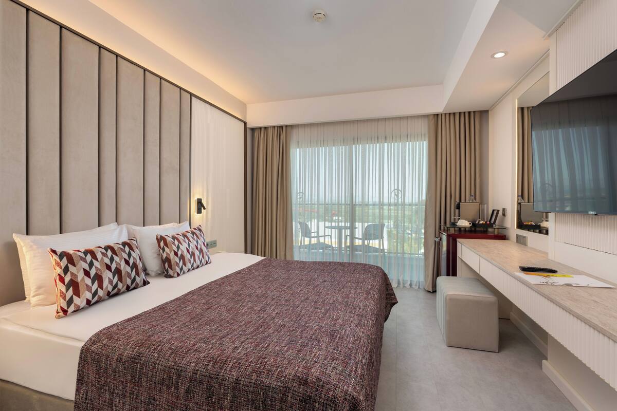 Ein modernes Hotelzimmer mit einem großen Bett, einem Schreibtisch, einem Flachbildfernseher und einem Balkon mit Sitzgelegenheit.