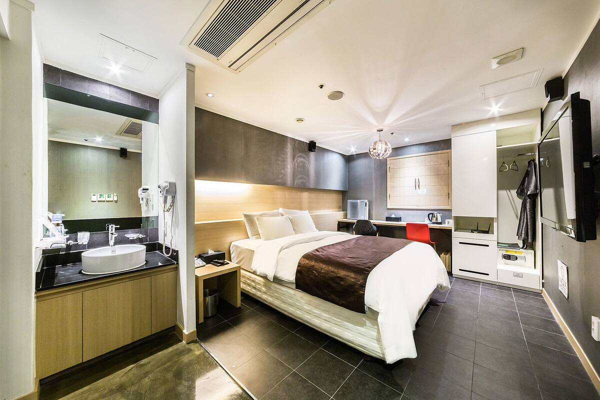 A modern hotel room with a large bed, a bathroom with a sink and mirror, a desk with a chair, and a wardrobe.