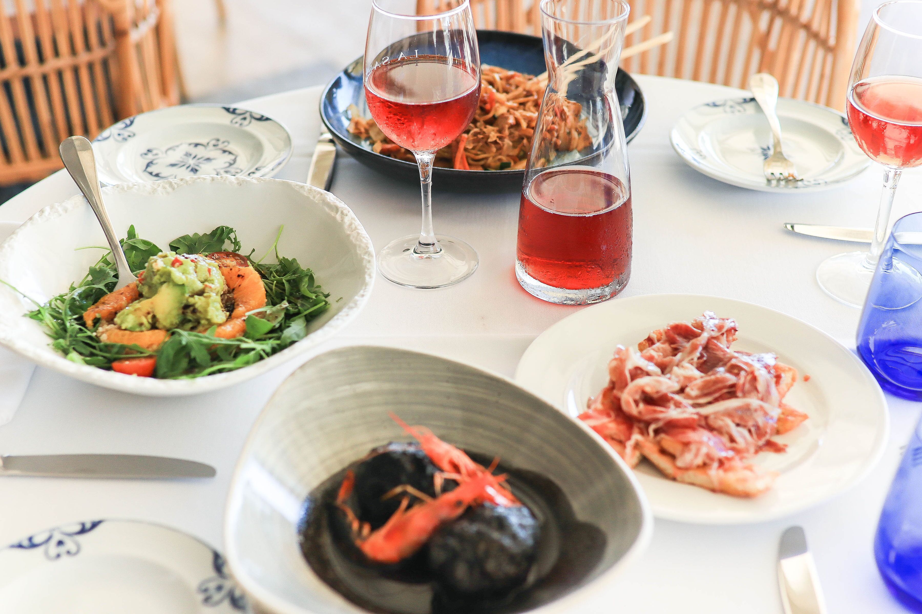 Une table dressée avec différents plats, notamment une salade, un bol de crevettes et une assiette de viande, ainsi que des verres de vin rouge.