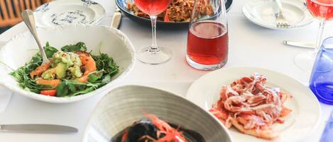Une table dressée avec différents plats, notamment une salade, un bol de crevettes et une assiette de viande, ainsi que des verres de vin rouge.
