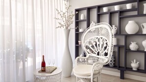 A white wicker chair with a cushioned seat and backrest, positioned next to a small round table with a bottle and two glasses. Behind the chair, a white vase with branches is on a pedestal, and a dark wooden shelf with various white ceramic items is visible. - Hotel Rosamar (Calonge)