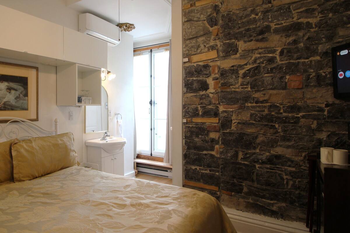 A bedroom with a bed, a bedside table, a mirror, a white sink, and a stone wall.