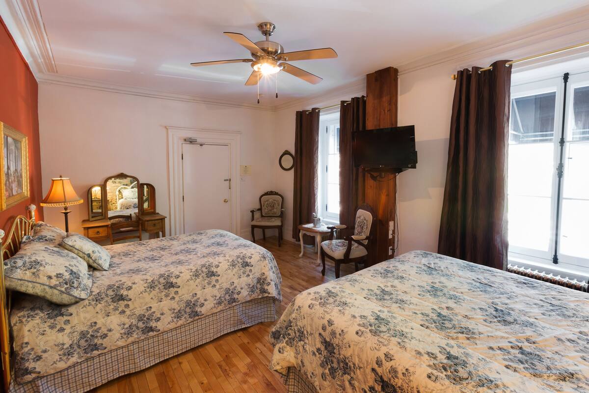 A hotel room with two beds, a ceiling fan, a TV, a desk, and a window with curtains.