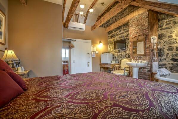 A bedroom with a large bed, a wooden ceiling, a stone wall, a white sink, and a mirror. - Manoir Sur le Cap (Québec City)