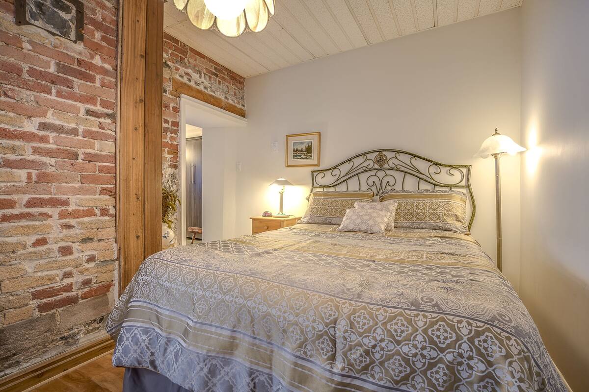A bedroom with a patterned bedspread, a brick wall, and a wooden ceiling.