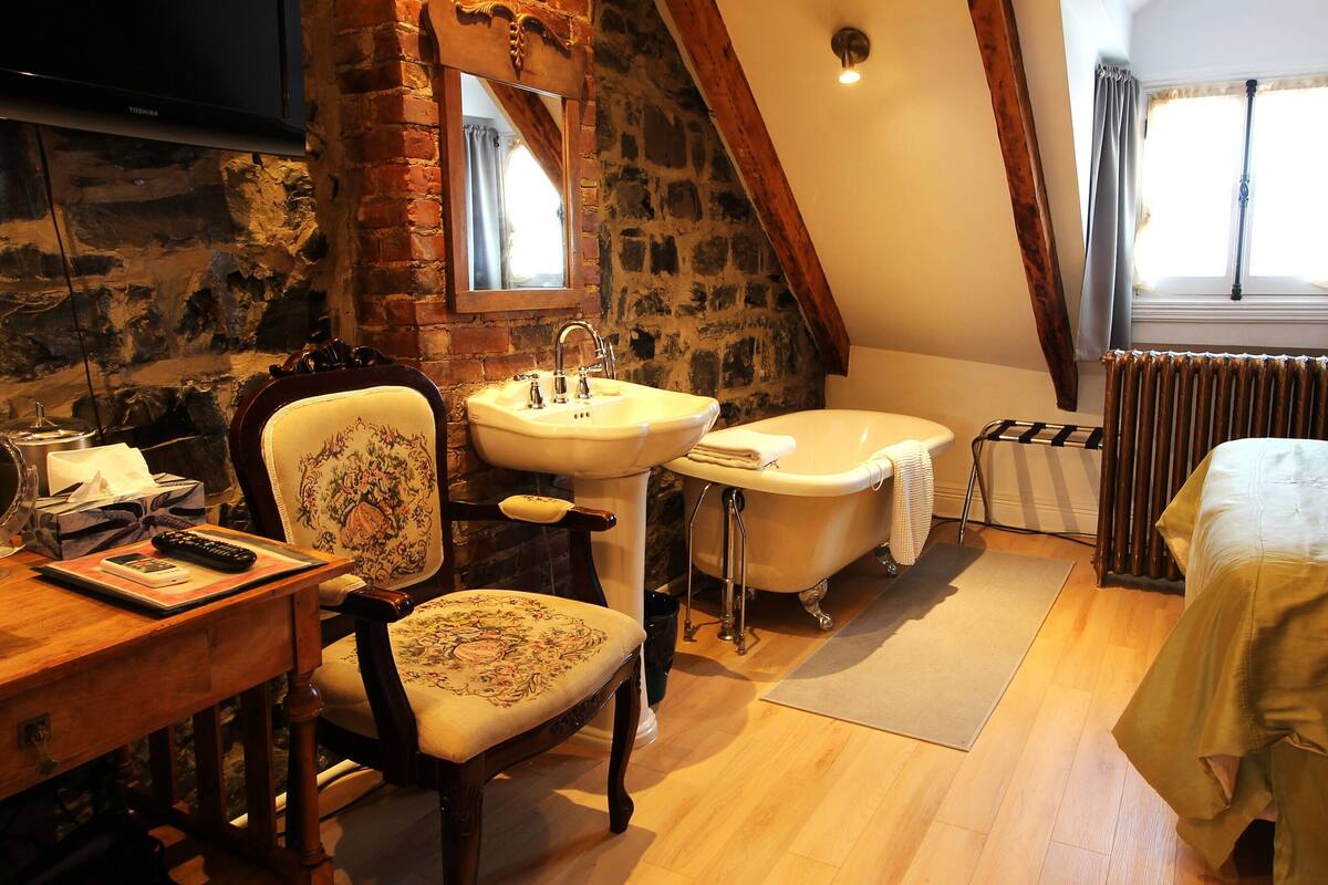 A room with a wooden chair, a wooden desk with a remote control, a white sink, a bathtub, a radiator, and a bed with a blanket.