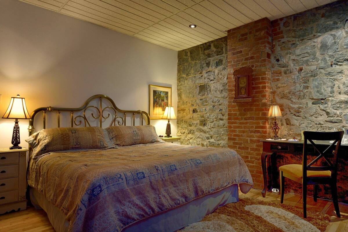 A bedroom with a stone wall, a bed with patterned bedding, two bedside lamps, a desk with a chair, and a wooden floor.
