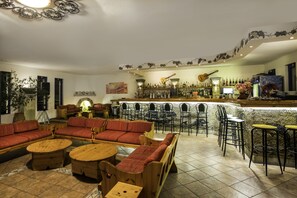 Eine Bar mit einer langen Theke, mehreren Barhocker und einer Auswahl an Flaschen. Es gibt verschiedene Sitzbereiche mit Sofas und Sesseln, und an der Wand hängt eine Gitarre.