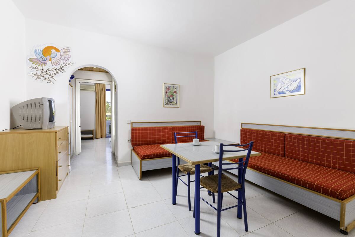 Une petite pièce propre, avec un bureau en bois, un banc rouge, une table avec des chaises et un téléviseur.