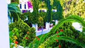 Un jardín exuberante con un edificio blanco, rodeado de follaje y flores rojas.