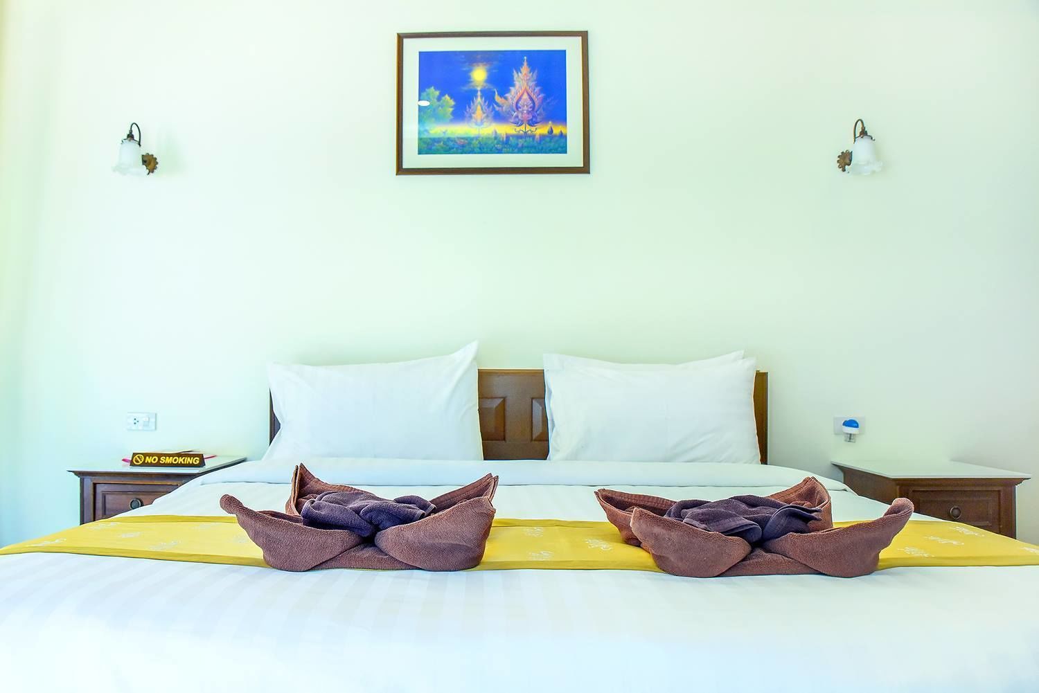 A hotel room with a bed, two pillows, a framed painting, and a 'No Smoking' sign.