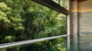 A modern indoor pool with a glass barrier, surrounded by a stone wall and a view of dense greenery.