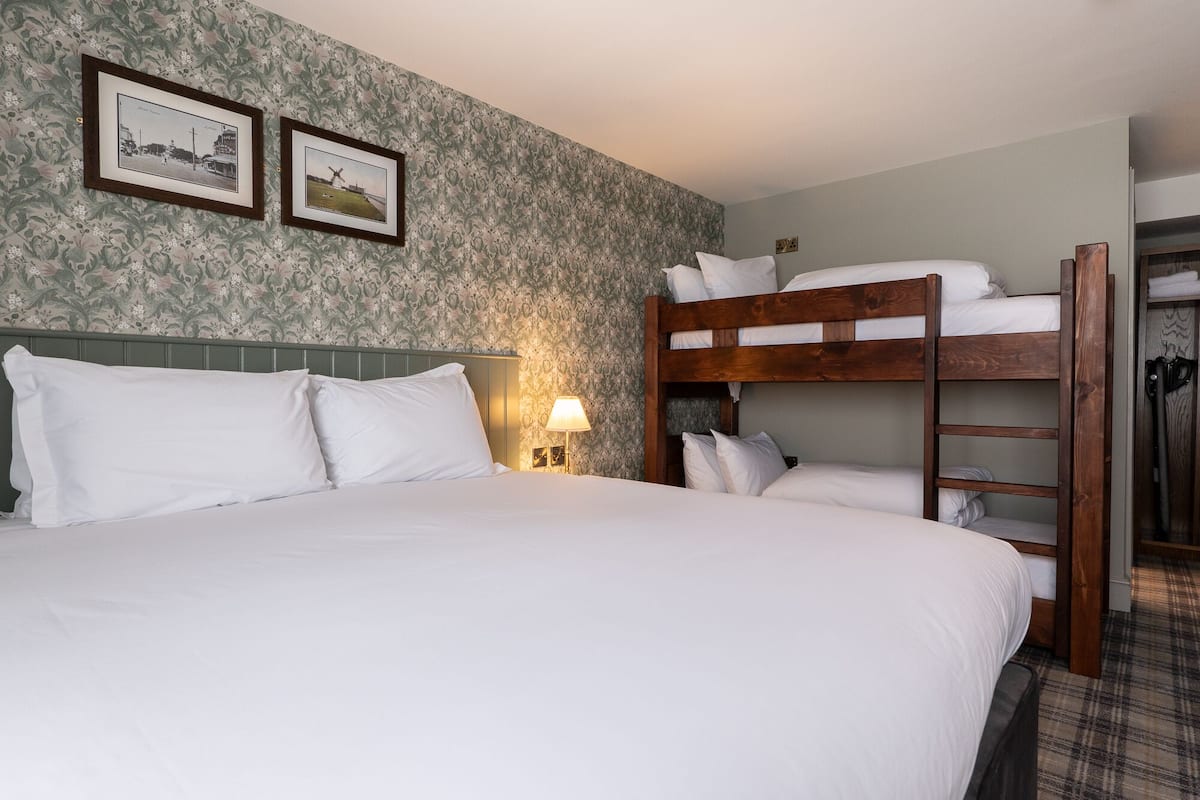 A hotel room with a bunk bed, a large bed with white linens, floral wallpaper, and two framed pictures on the wall.