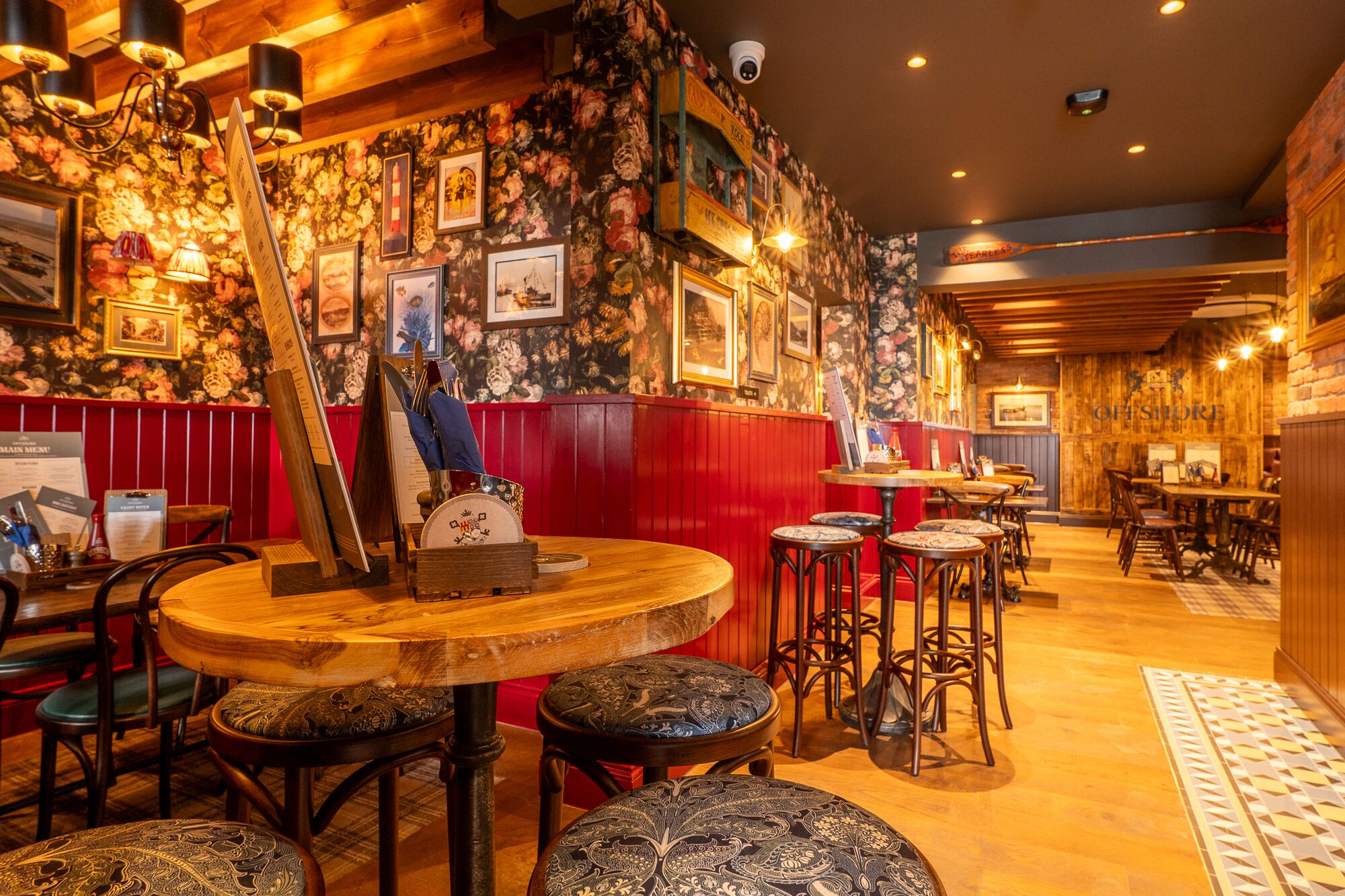 A cozy restaurant interior with wooden tables and chairs, floral wallpaper, and a red accent wall.