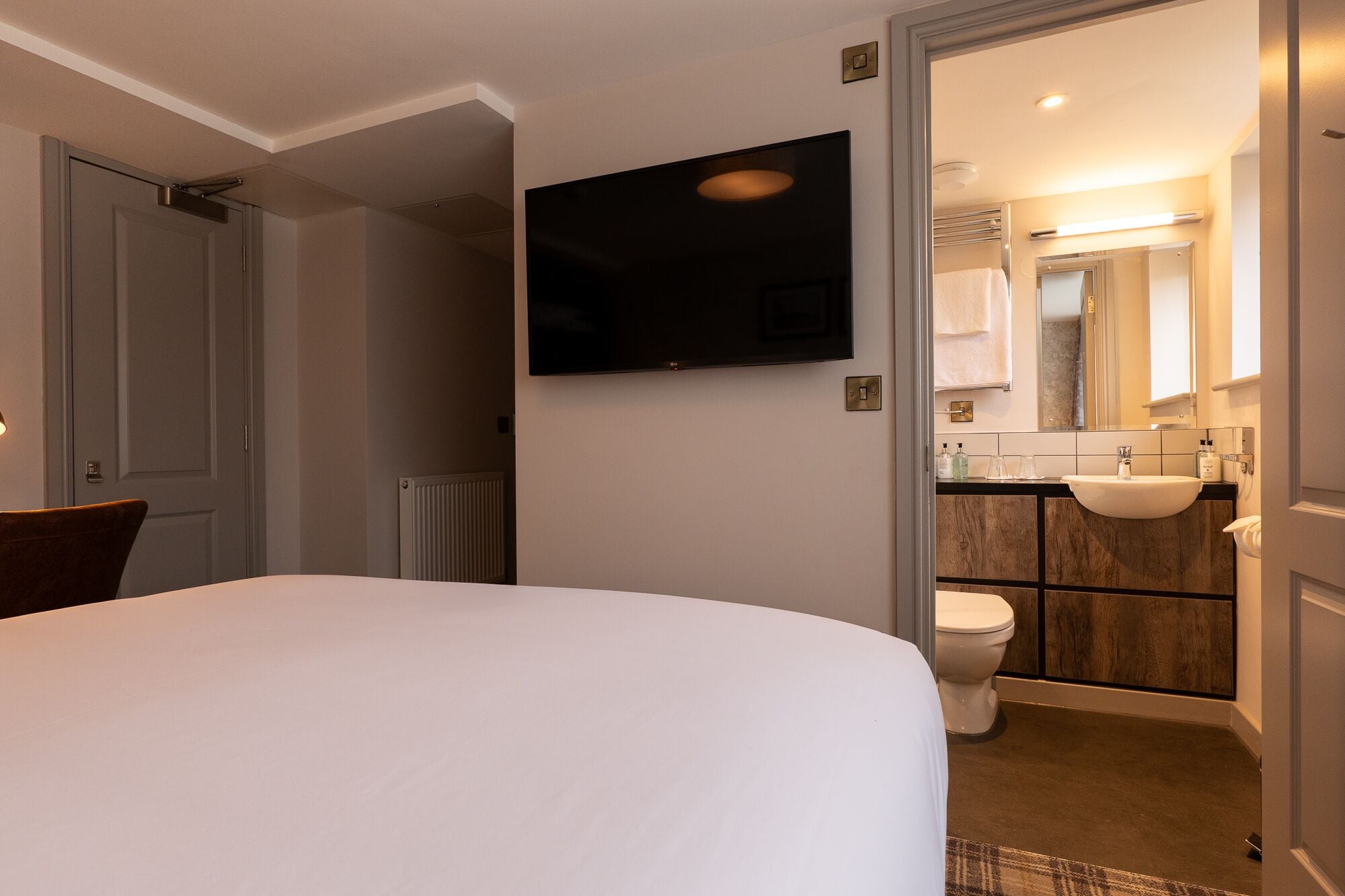 A hotel room with a bed, a TV mounted on the wall, a bathroom with a sink and toilet, and a radiator.