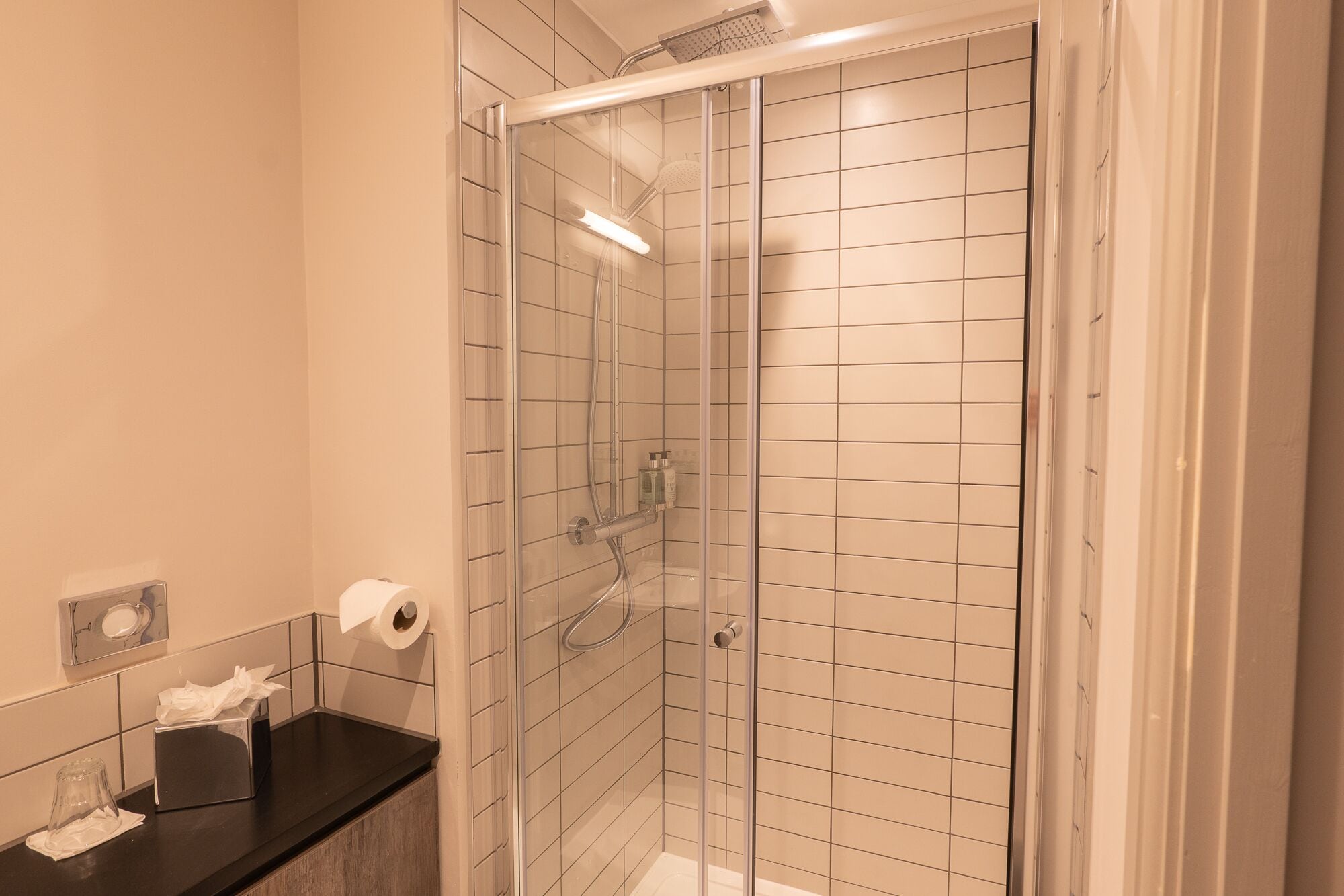 A modern bathroom with a glass-enclosed shower, white tiled walls, and a black countertop with toiletries.