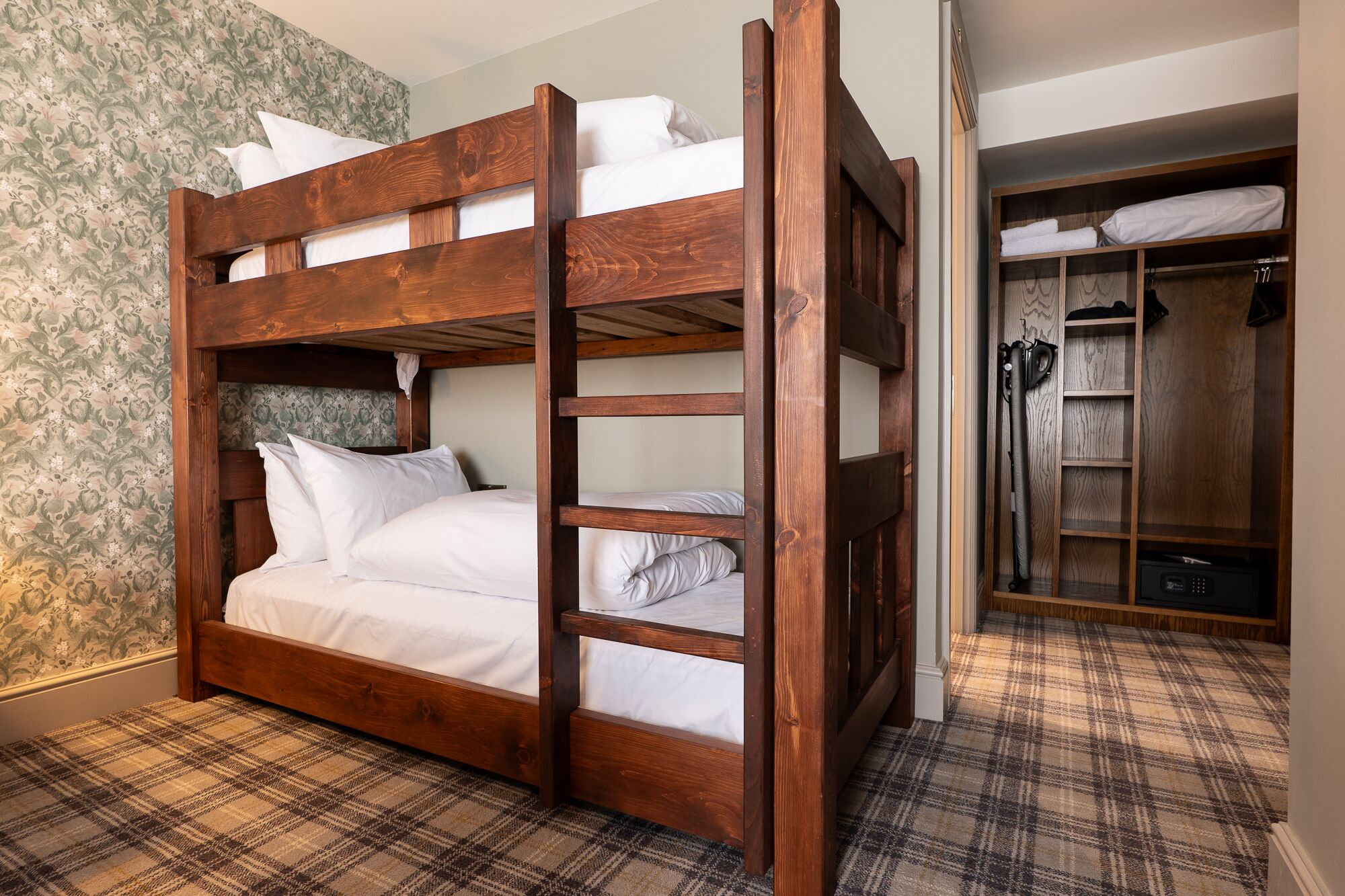A bunk bed with a wooden frame, white bedding, and a plaid carpet. There is a closet with a mirror and a hanging rack.