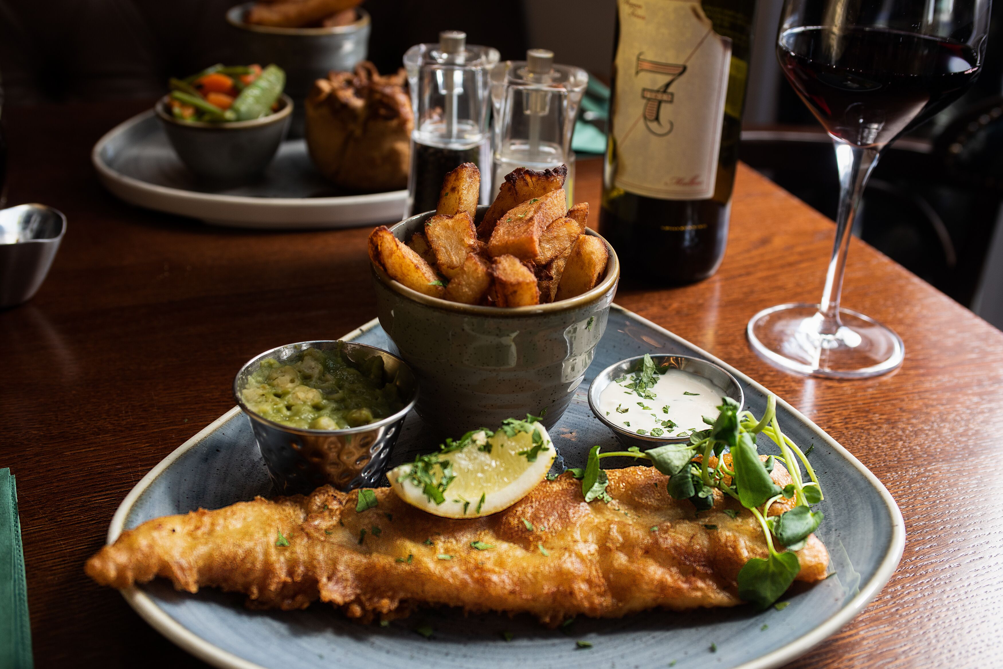 A plate of fish and chips with sides of mushy peas and tartar sauce, a glass of red wine, and a bottle of wine on a wooden table.