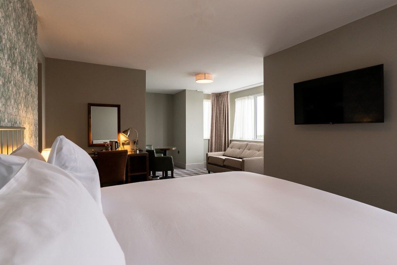 A hotel room with a large bed, a desk with a lamp, a sofa, a TV mounted on the wall, and a window with curtains.