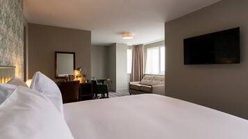 A hotel room with a large bed, a desk with a lamp, a sofa, a TV mounted on the wall, and a window with curtains.
