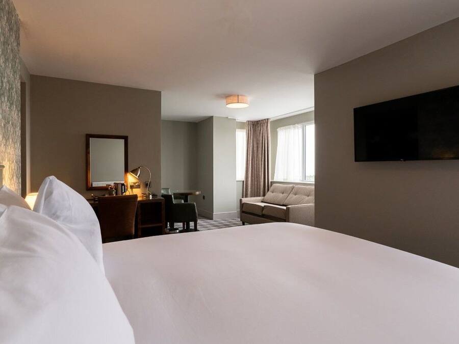 A hotel room with a large bed, a desk with a lamp, a sofa, a TV mounted on the wall, and a window with curtains.