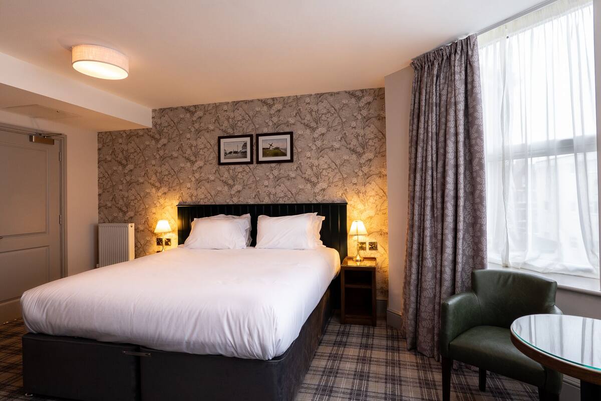 A hotel room with a large bed, two bedside lamps, a small table, a chair, and a window with curtains.