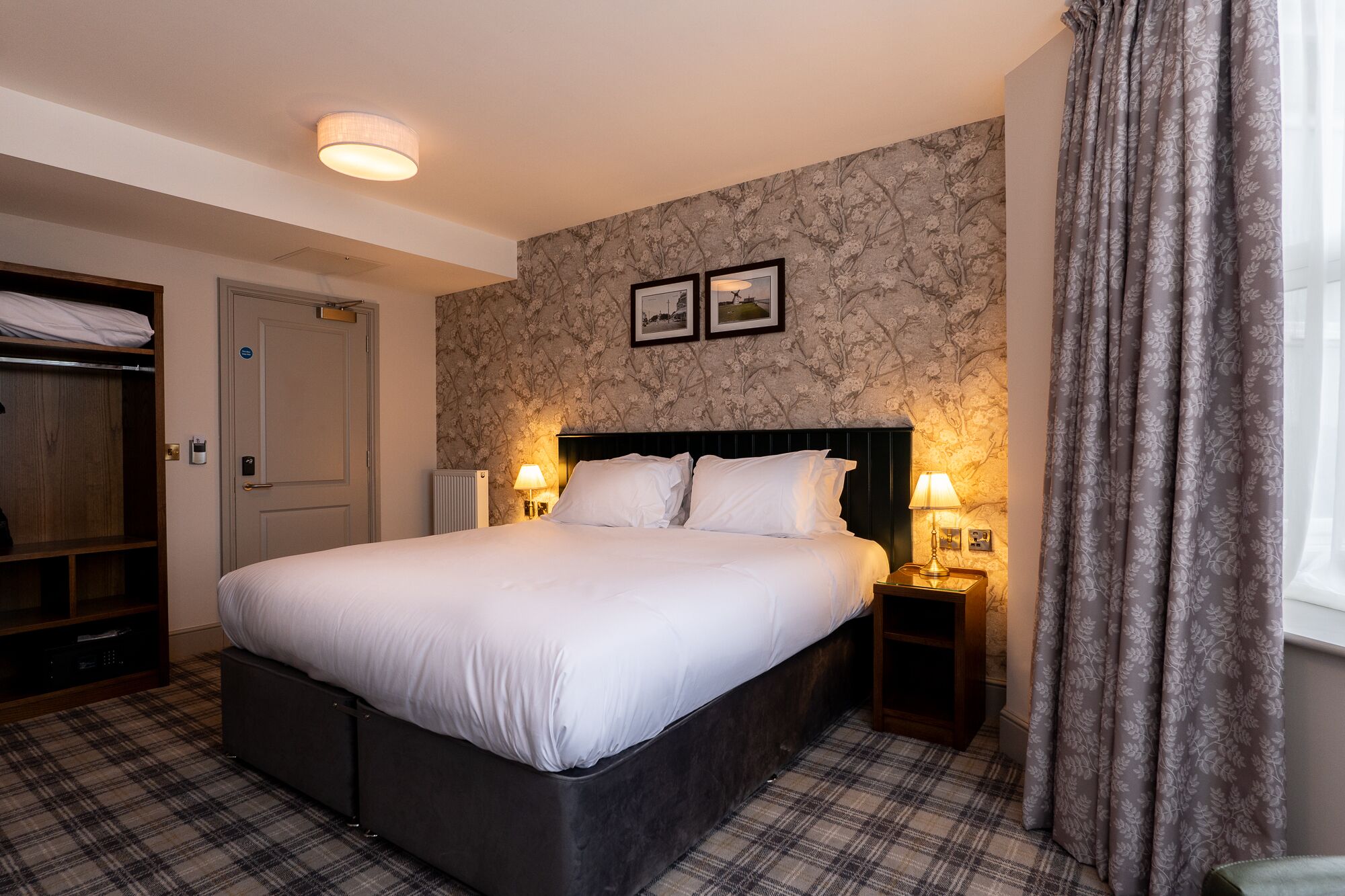 A hotel room with a large bed, two bedside lamps, a wardrobe, and a floral-patterned wall.