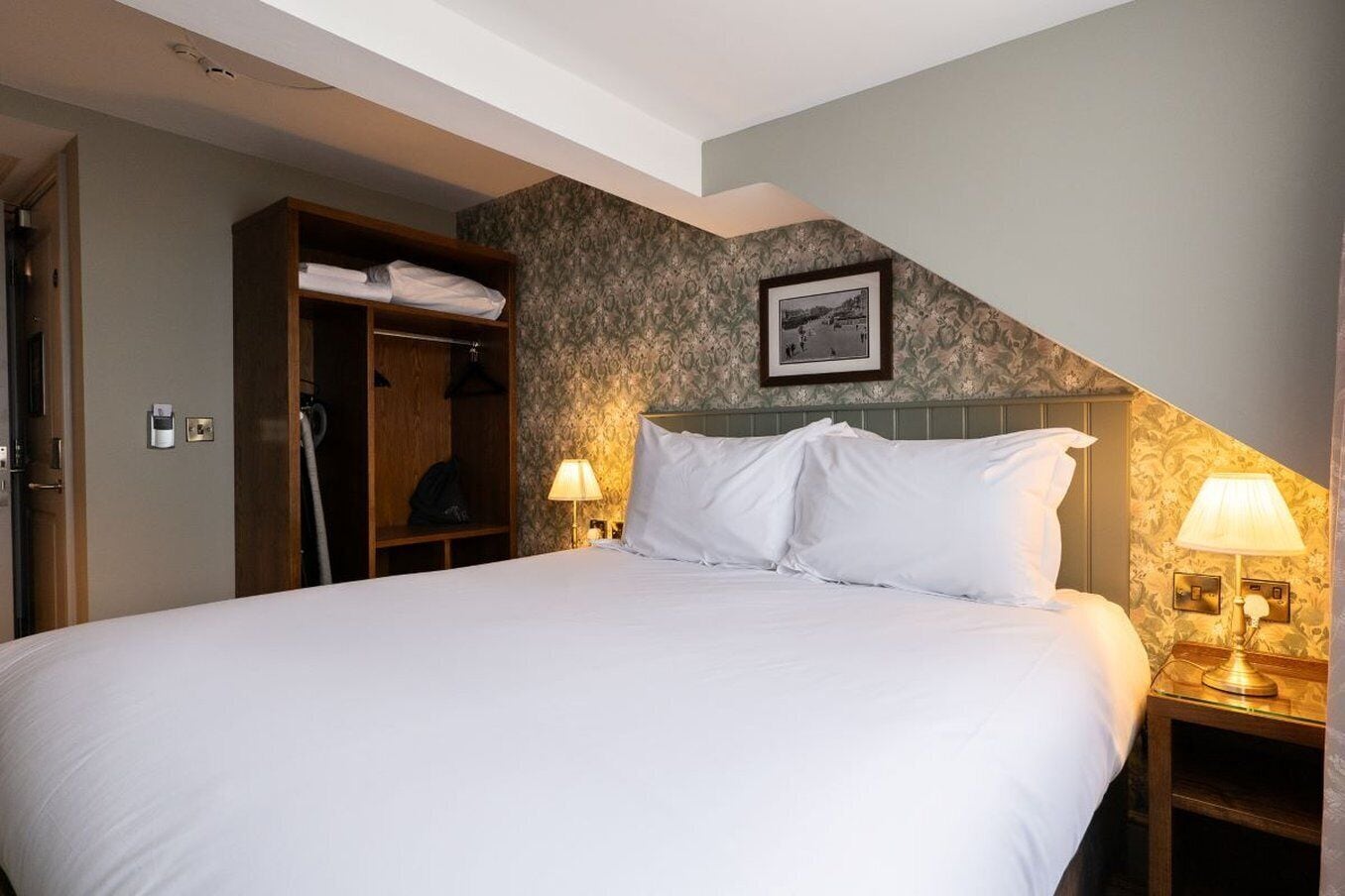 Una cama bien hecha en una habitación de hotel, con un armario de madera, dos lámparas de noche y un cuadro en la pared.