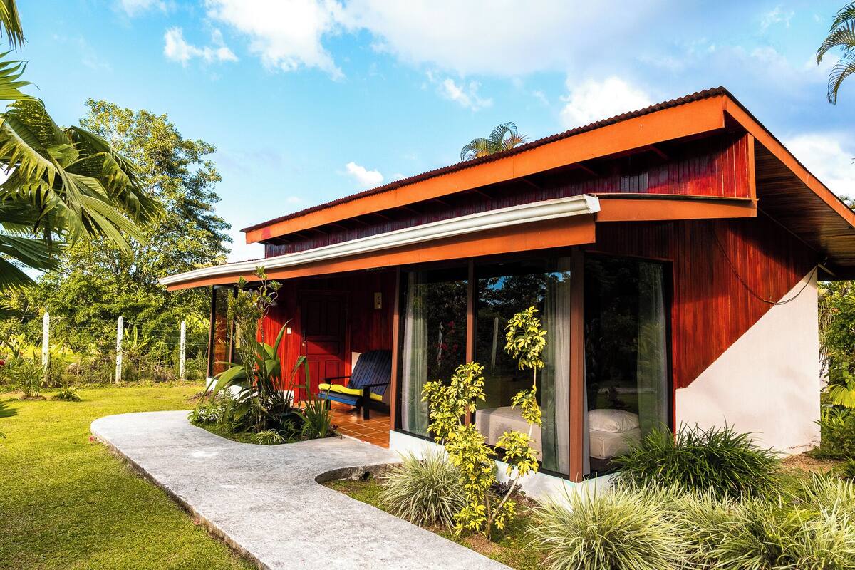 A modern house with a red roof, large glass windows, and a concrete pathway leading to the entrance.