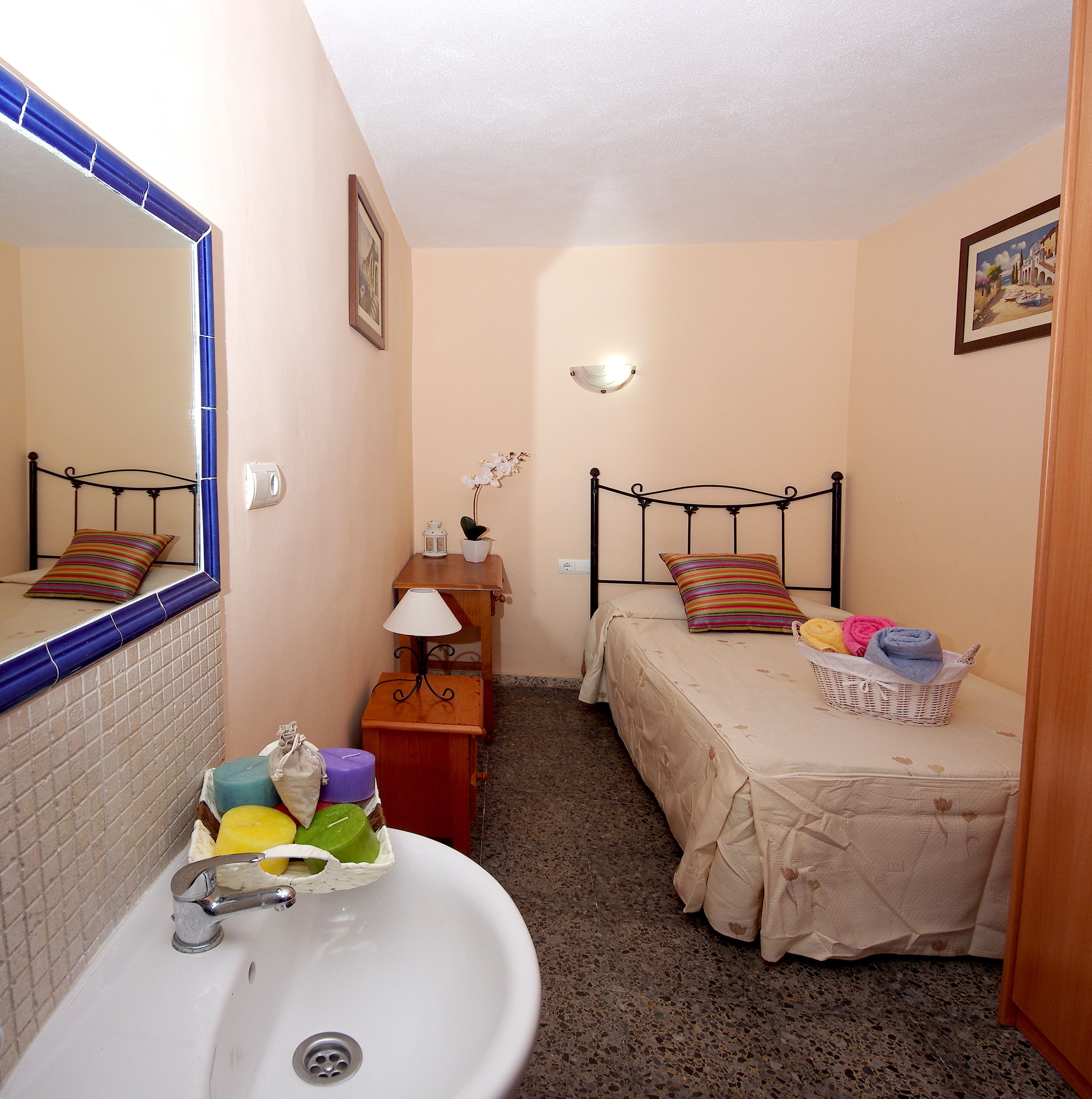 A small, well-lit bedroom with a single bed, a bedside table with a lamp, a mirror, and a window with a view of the outside.