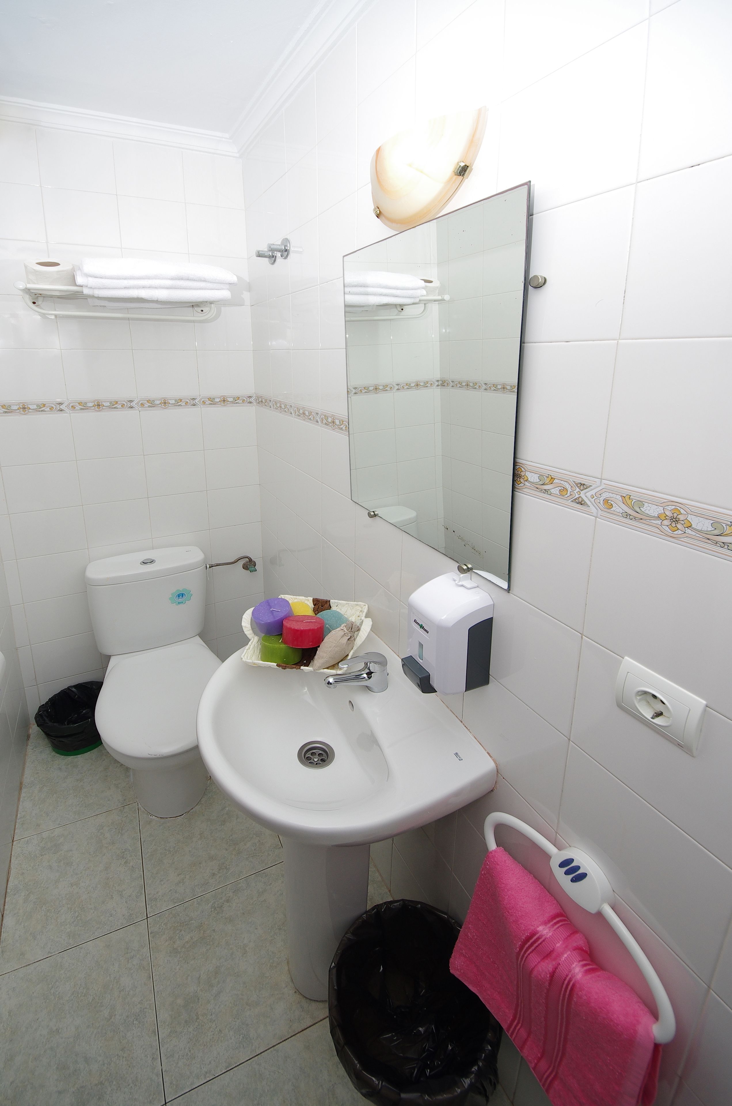 A bathroom with a toilet, sink, and mirror. There is a trash bin and a towel rack with a pink towel.