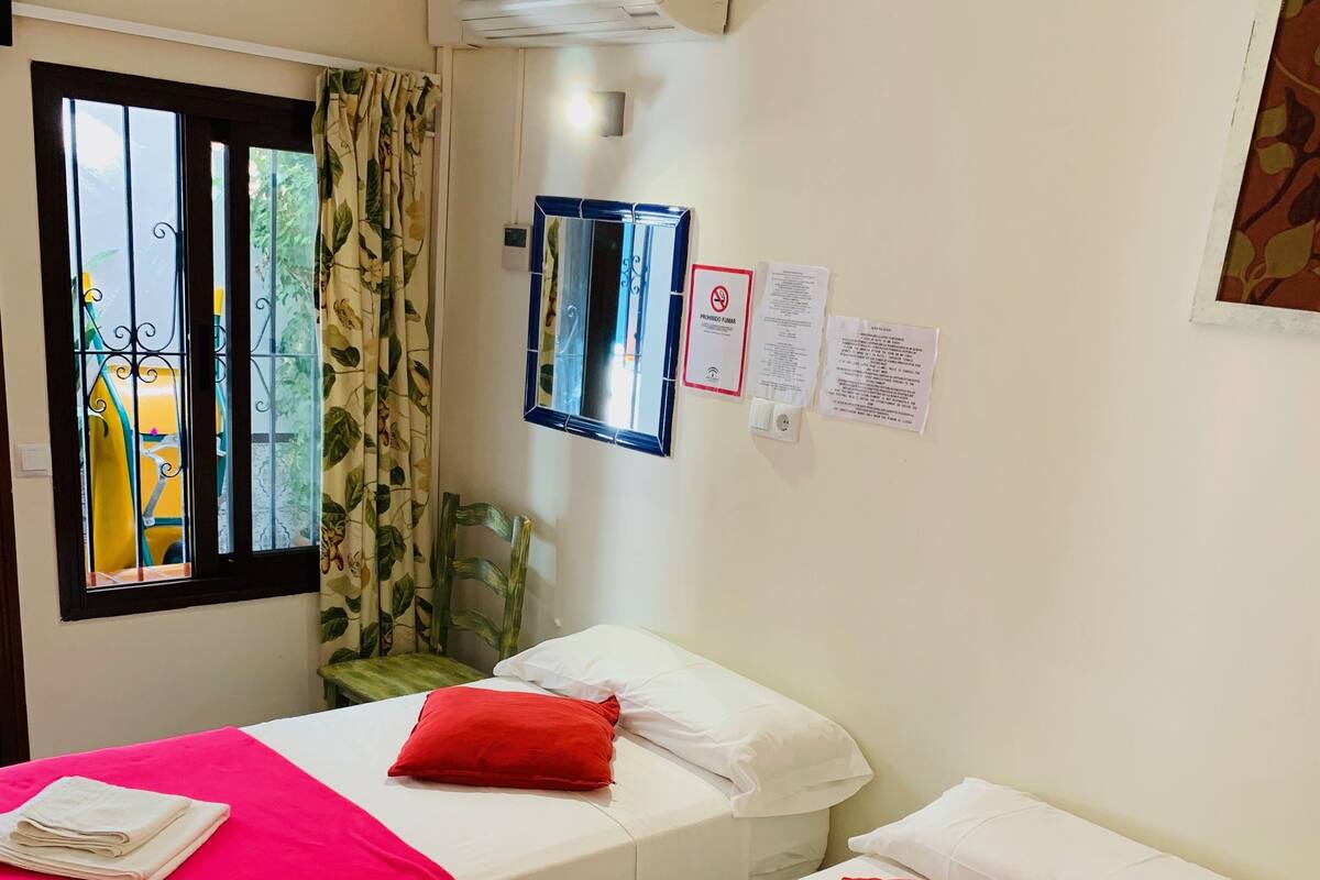 A hotel room with two beds, each with a red and white bedspread, and a small table with a folded towel. There is a window with a view of greenery outside, an air conditioning unit, and a chair.