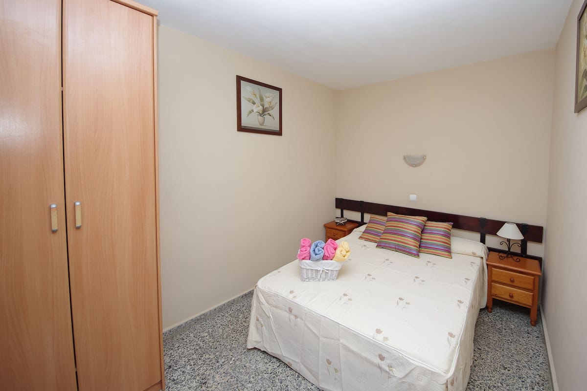 A bedroom with a bed, a nightstand, a bedside table, a wooden wardrobe, and a wall-mounted picture.