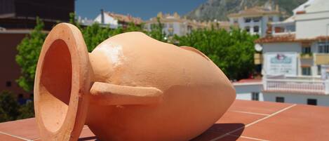 Una maceta de terracota sobre una superficie de baldosas, con edificios y vegetación al fondo.