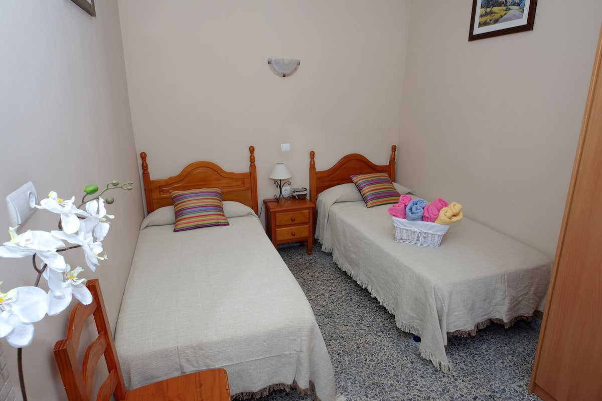 A bedroom with two wooden beds, a nightstand with a lamp, and a small table with a basket of towels.
