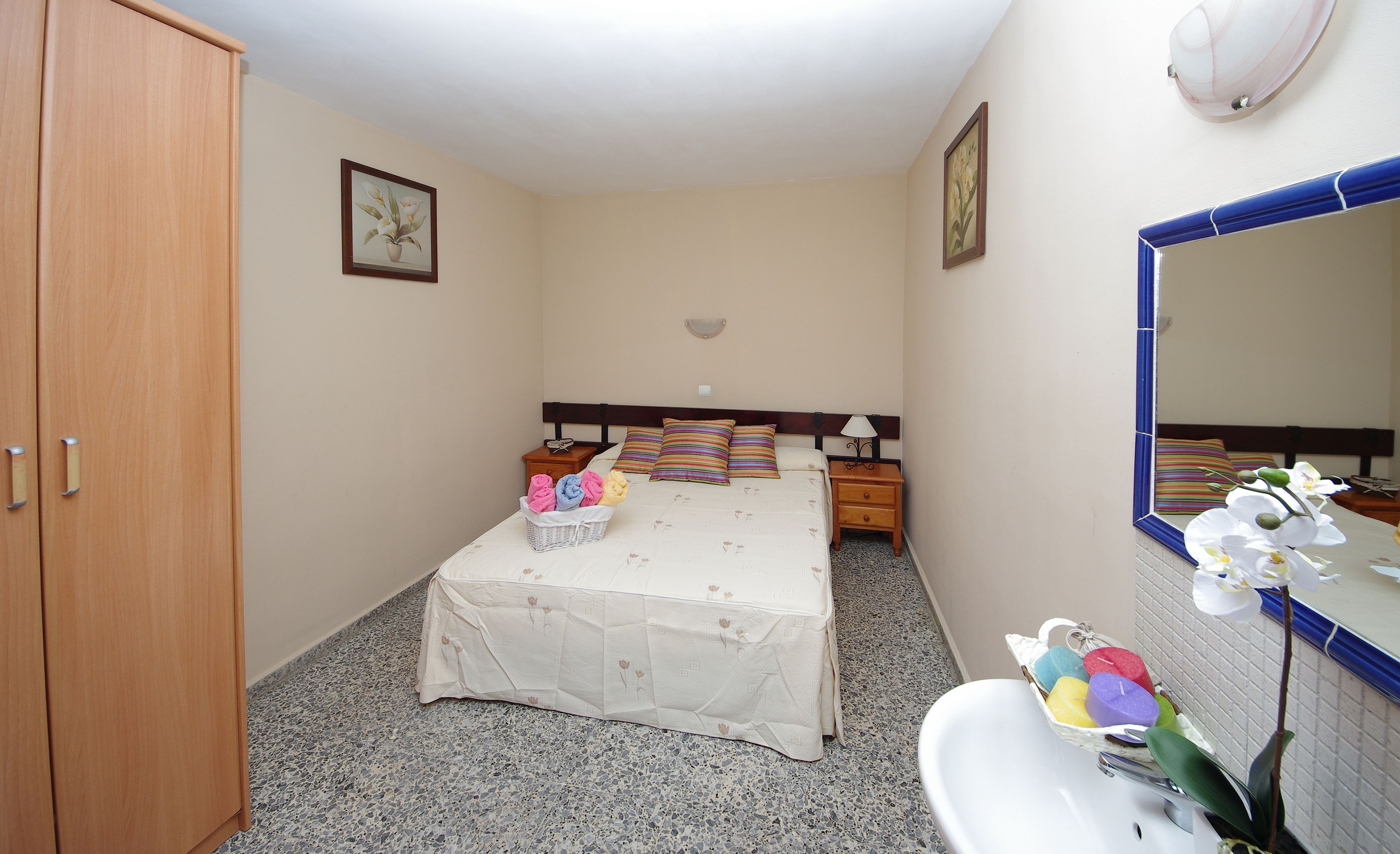 Een slaapkamer met een bed, een kledingkast, een nachtkastje, een lamp, een spiegel en een badkamer met een wastafel en een bloevas.