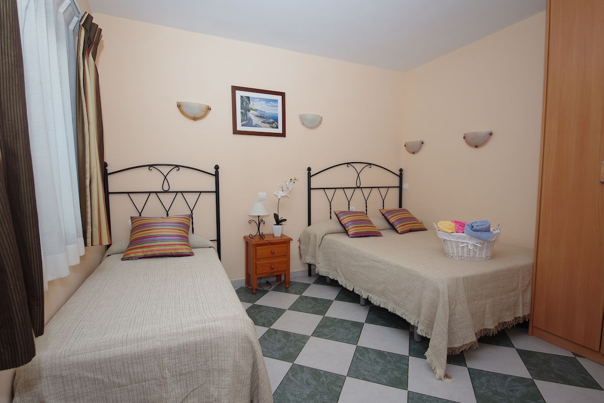 A bedroom with two single beds, a nightstand with a lamp, a framed picture on the wall, and a checkered floor.