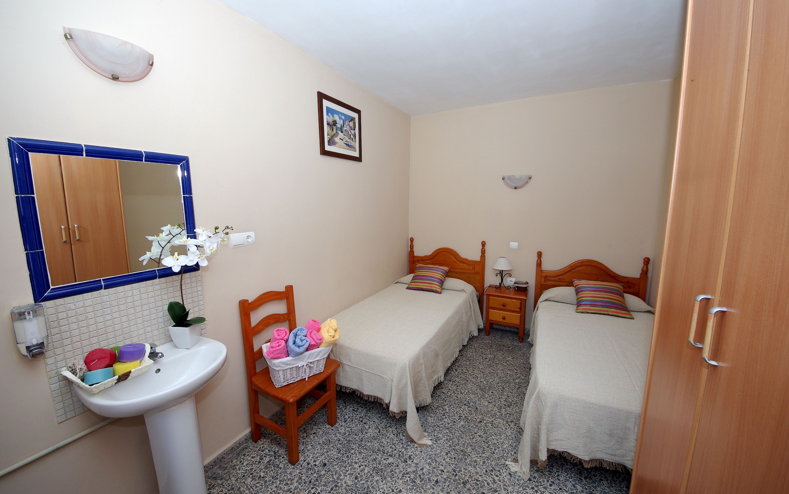 A small, clean room with two single beds, a wooden chair, a white sink, and a mirror above the sink.