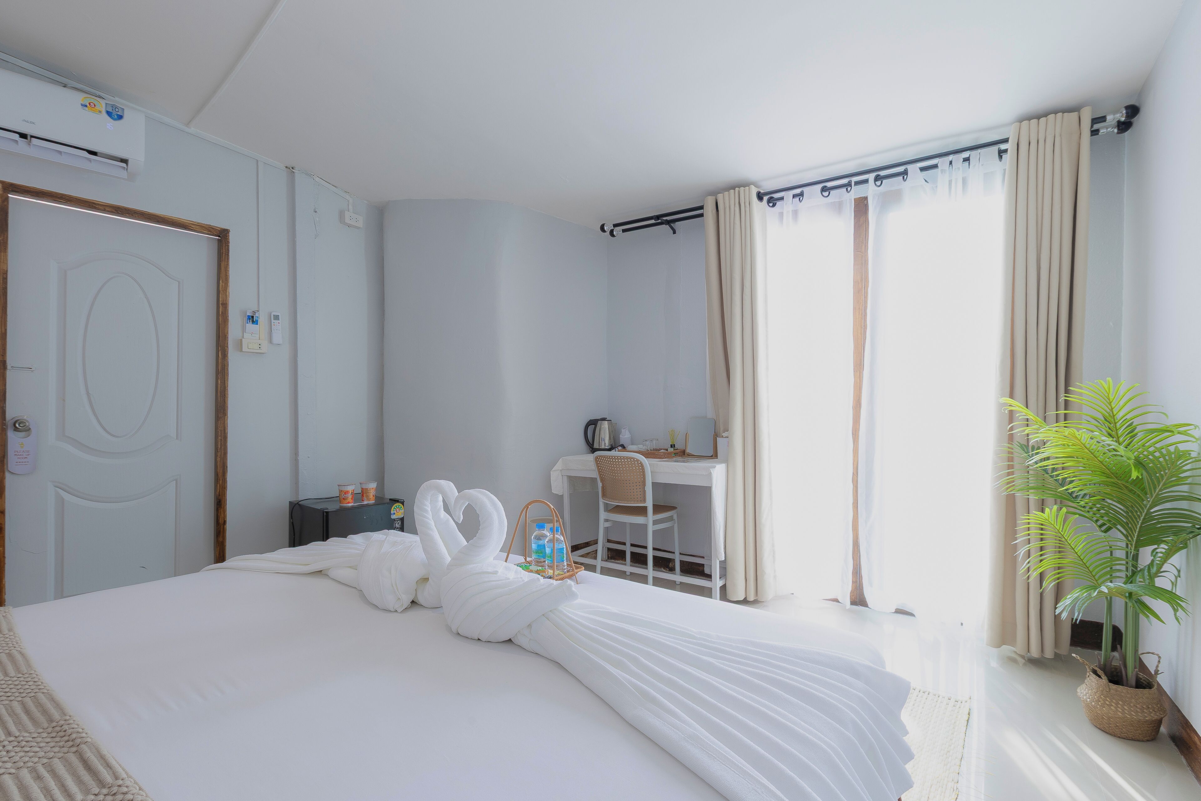 A hotel room with a bed, a desk, a chair, a potted plant, and a window with curtains.