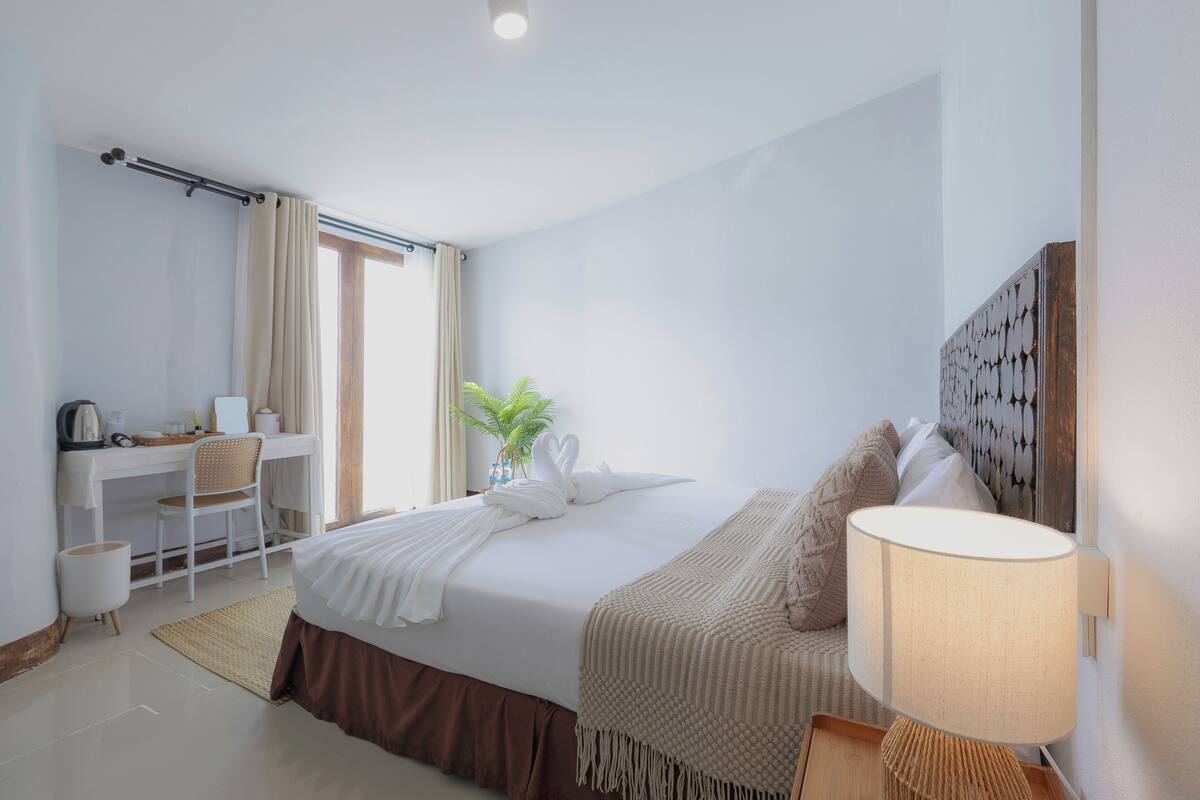 Habitación de hotel con cama, escritorio, silla, lámpara y ventana con cortinas.