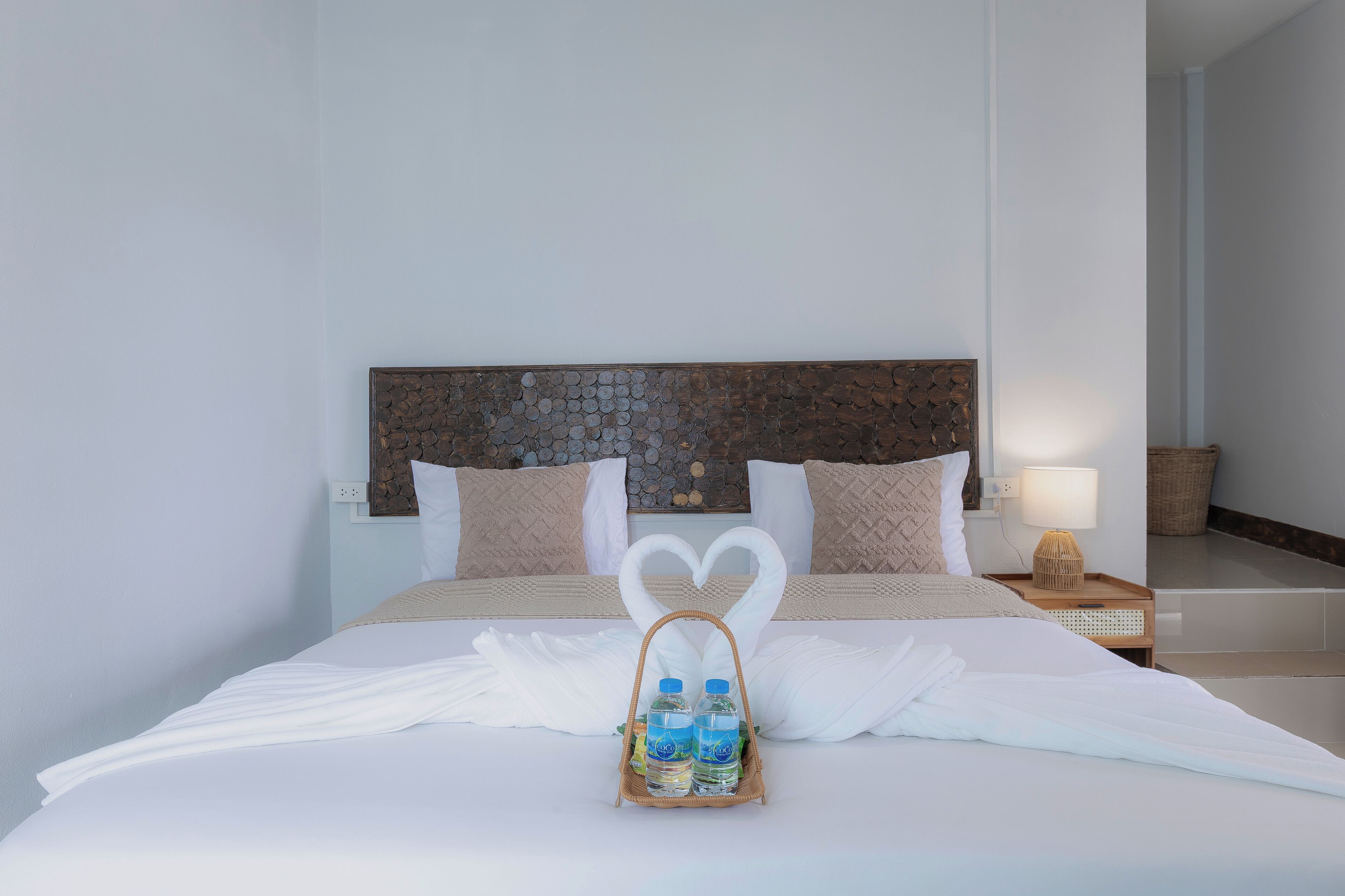 Habitación de hotel con cama, ropa de cama blanca, cabecera con diseño de mosaico, dos almohadas, mesita de noche con lámpara y bandeja con botellas de agua.