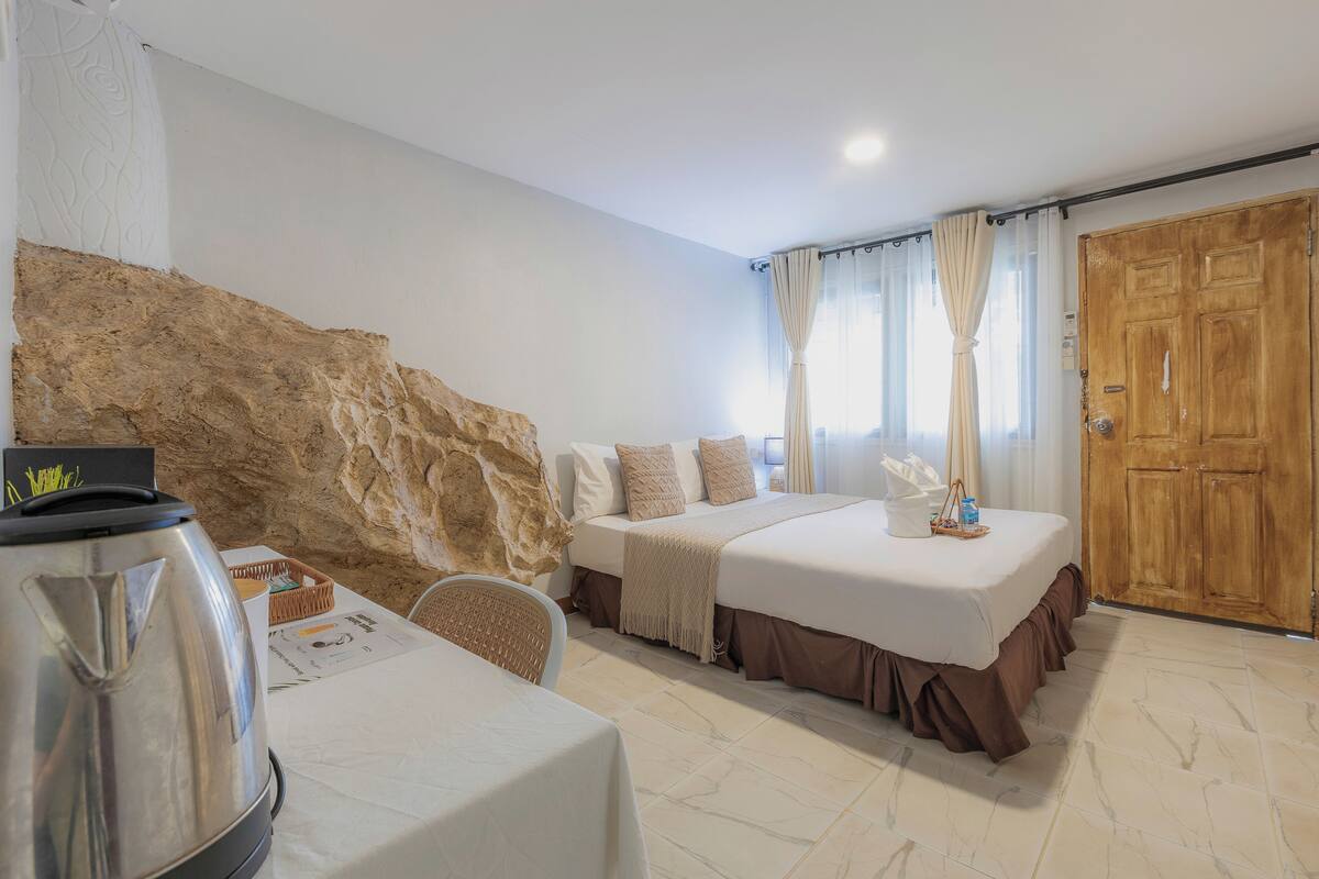 Habitación de hotel con una gran pared de roca, una cama con falda marrón, una puerta de madera y una mesa con tetera.