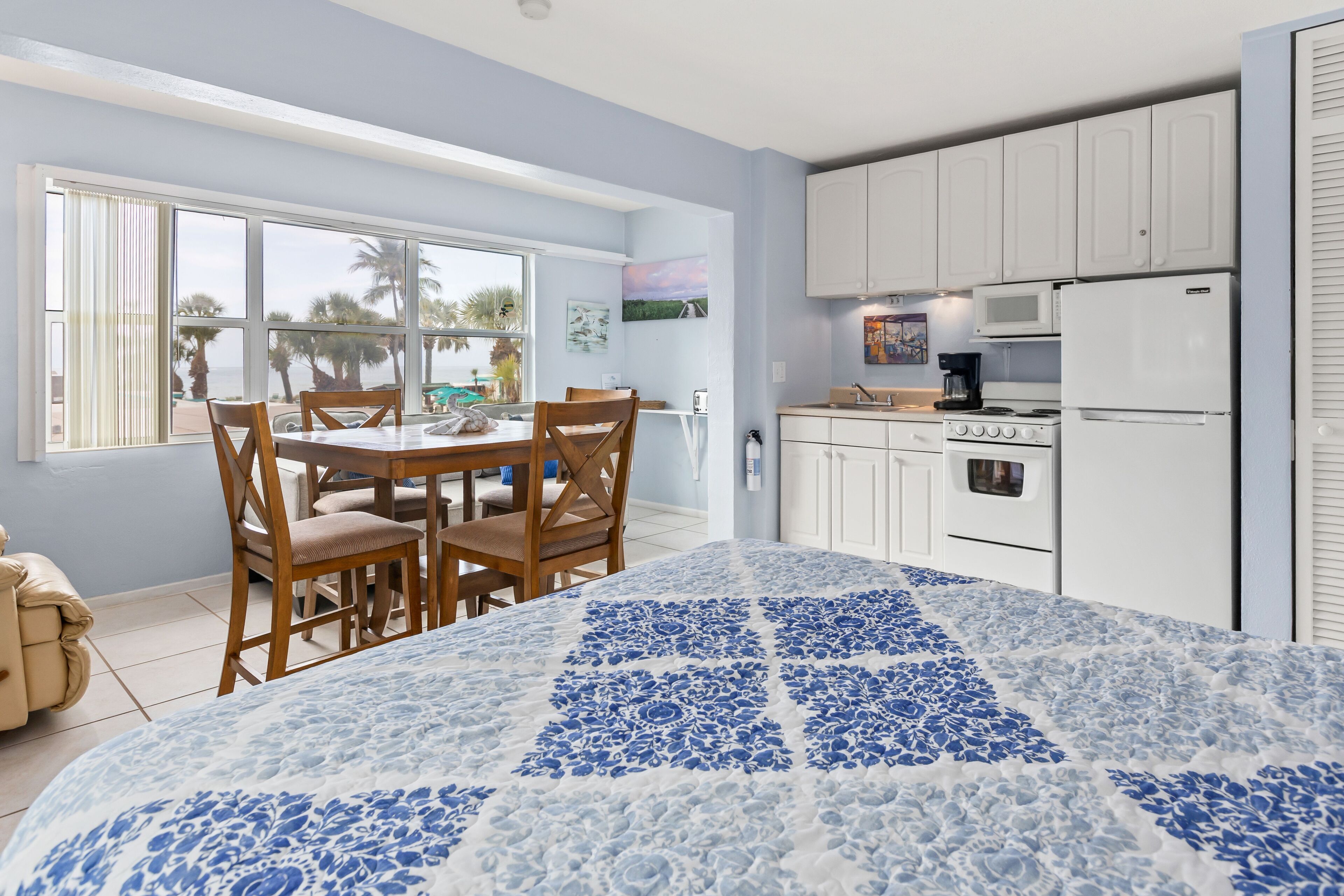 A dining area with a table and chairs, a kitchen with white cabinets and appliances, and a view of palm trees through large windows.