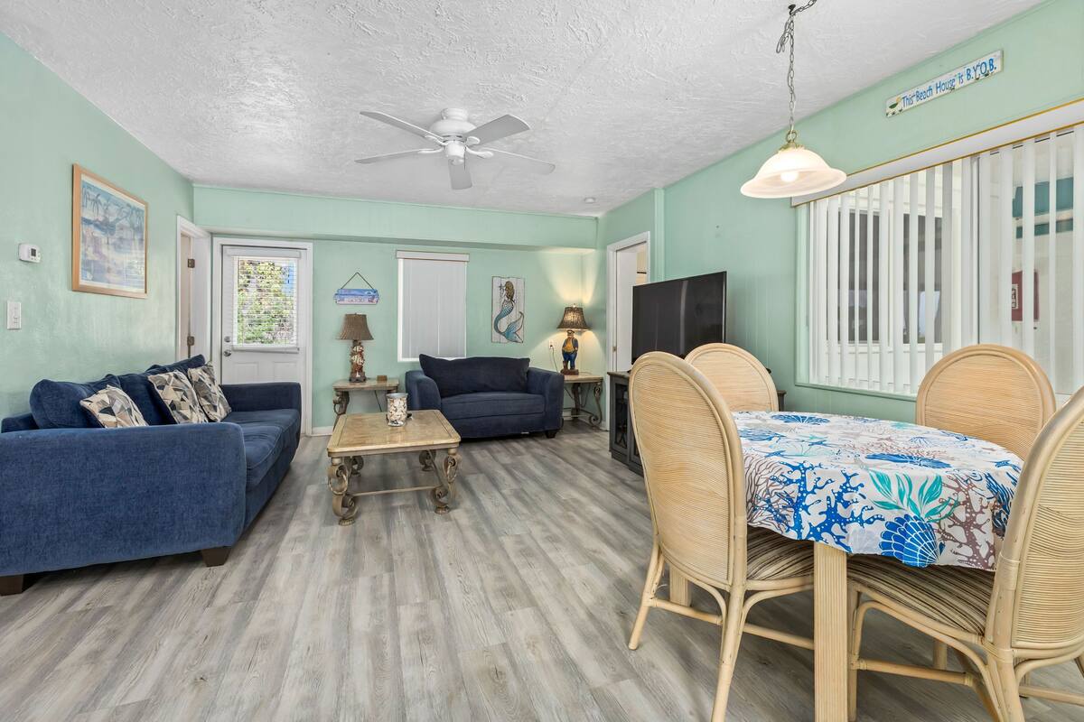 Ein Wohnzimmer mit blauem Sofa, einem Holztisch, Korbstühlen, einem Esstisch mit floralem Tischläufer, einem Deckenventilator und einem Fernseher an der Wand.