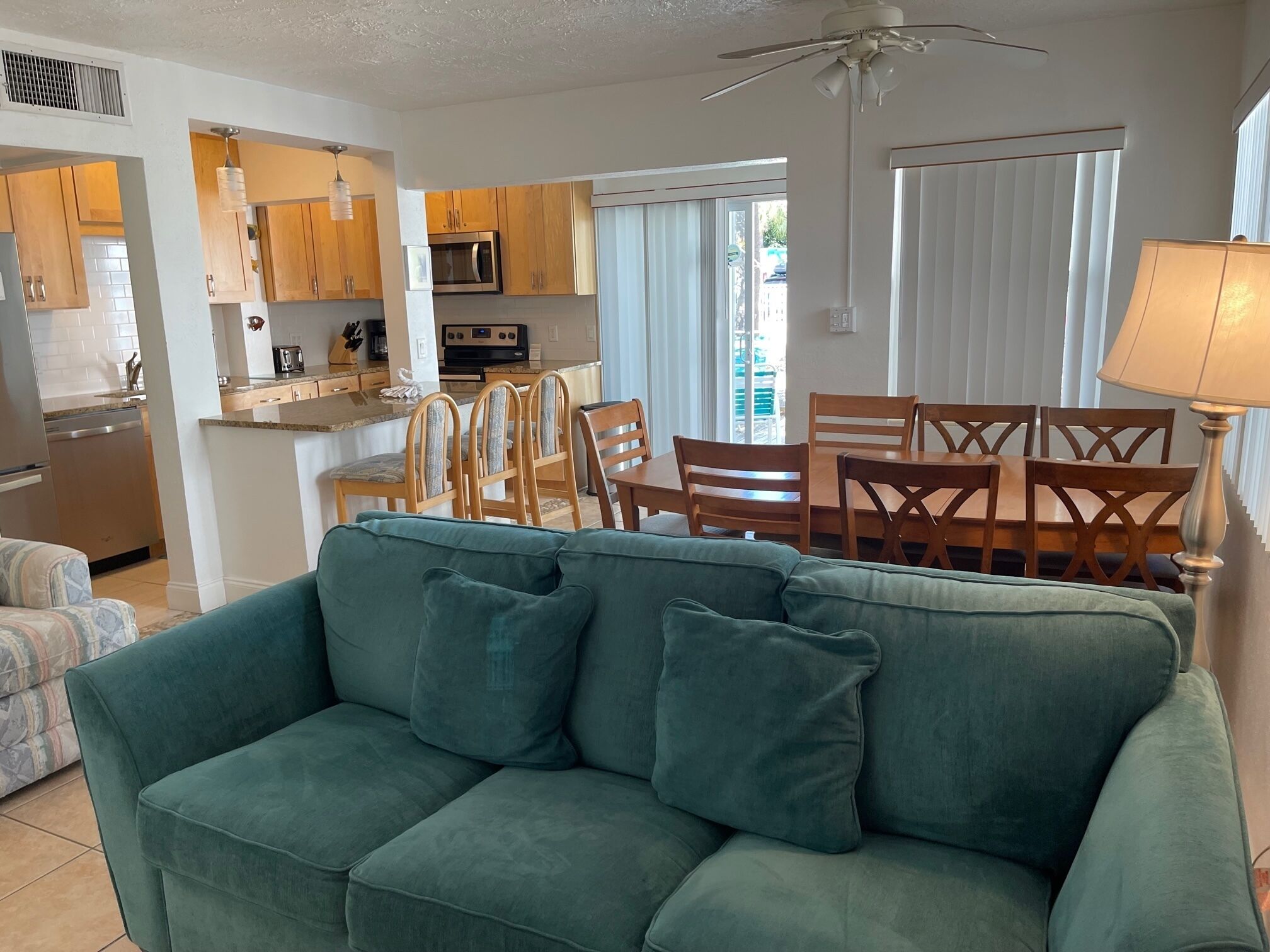 A modern living room with a green sofa, wooden dining table, and chairs. A kitchen with wooden cabinets and appliances is visible in the background.