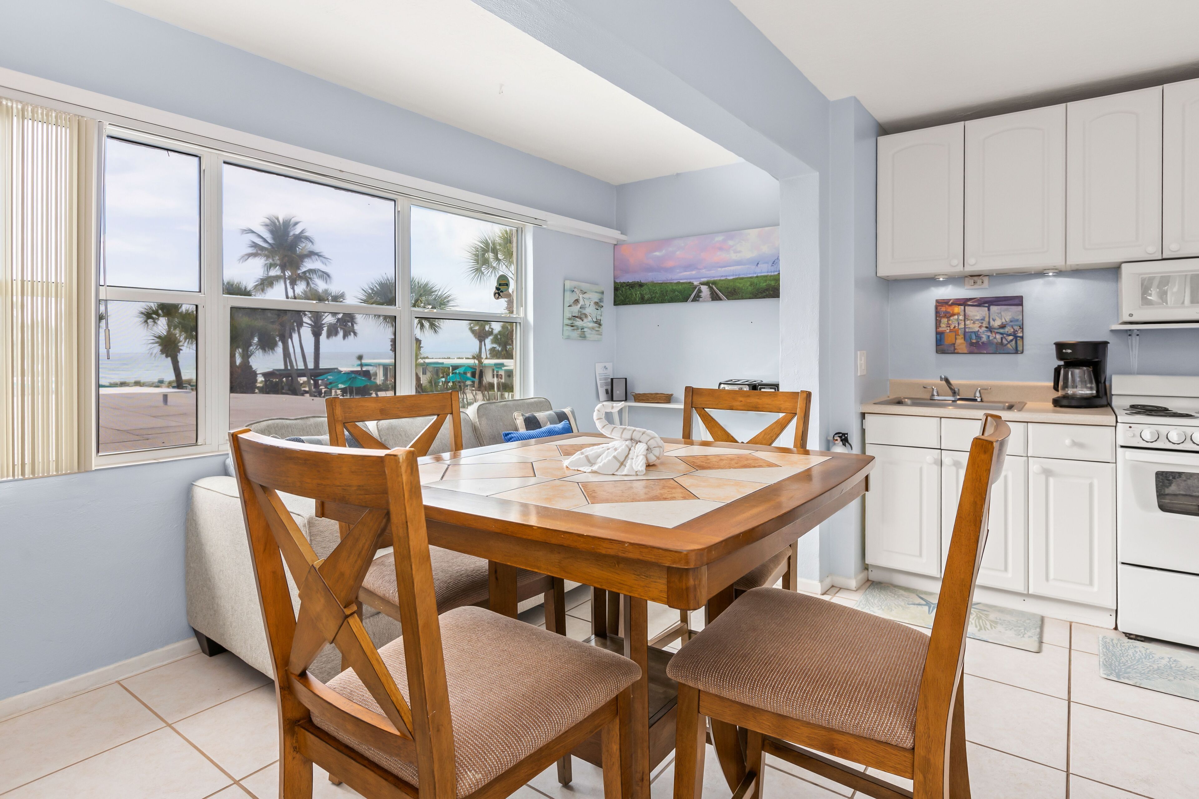 Un coin repas avec une table et des chaises en bois, une cuisine avec des meubles blancs et une vue sur des palmiers et une plage, le tout visible à travers de grandes fenêtres.