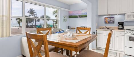 Un coin repas avec une table et des chaises en bois, une cuisine avec des meubles blancs et une vue sur des palmiers et une plage, le tout visible à travers de grandes fenêtres.