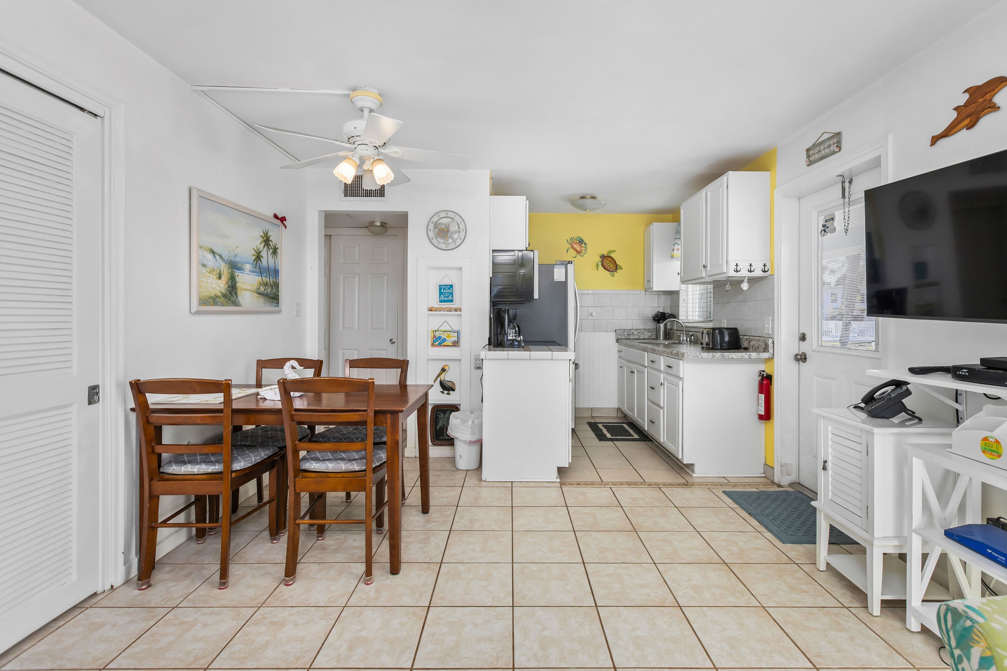 A compact kitchen with white cabinets, a refrigerator, and a microwave. There is a dining table with chairs and a ceiling fan.